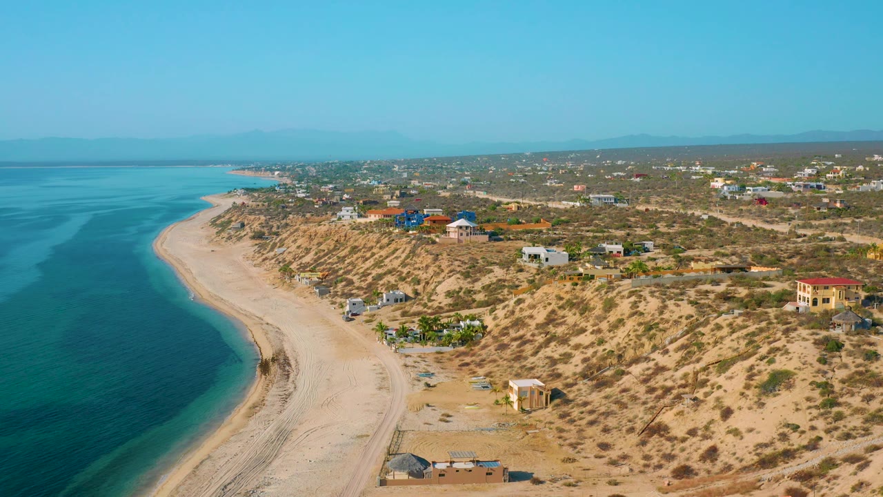 Wide aerial frame from El Sargento La Ventana, Mexico, with the sea occupying the left side, pale beach below, dry scrubland and small houses to the right, and no visible people.