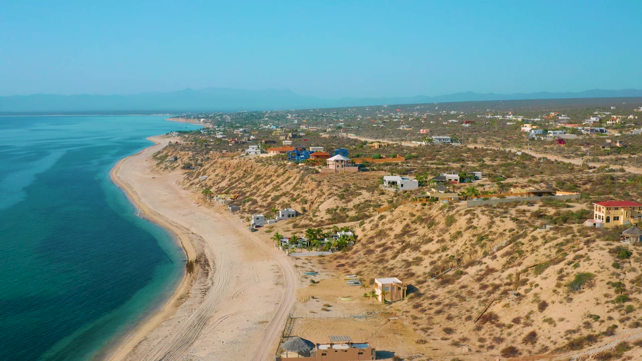 Aerial beach curve with calm sea, sandy dunes, houses, and distant mountains.