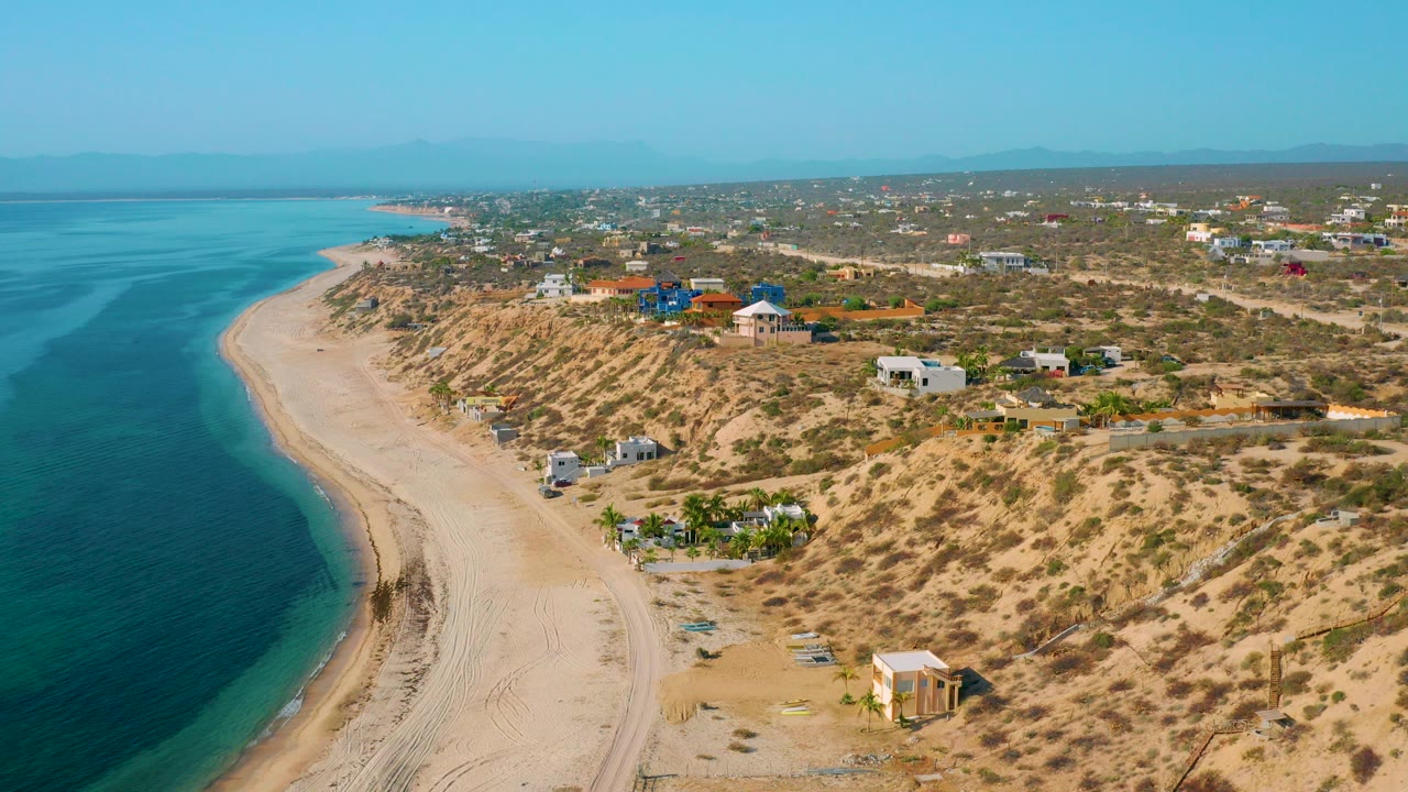 Elevated view of El Sargento La Ventana, Mexico, with turquoise water on the left, a pale sandy beach curving below, scrub dunes, scattered houses, and no visible people.