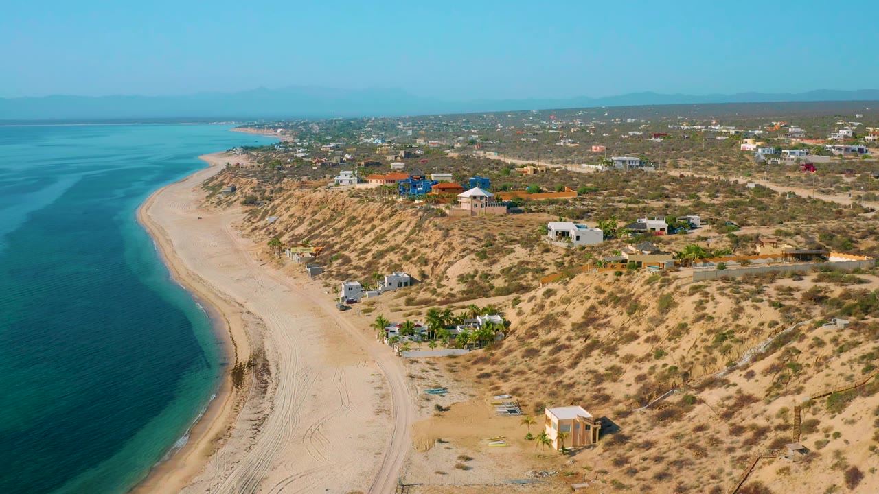 Aerial frame over El Sargento La Ventana, Mexico, showing calm blue-green water beside a long sand strip, dry vegetation, small buildings, and no visible people.