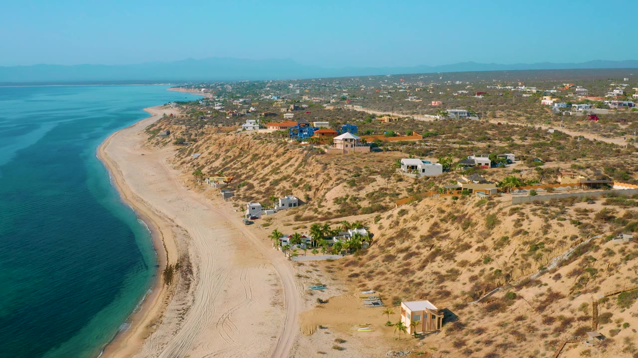 Drone-height view of El Sargento La Ventana, Mexico, with the sea filling the left side, a broad sandy beach, desert scrub, inland homes, and no visible people.