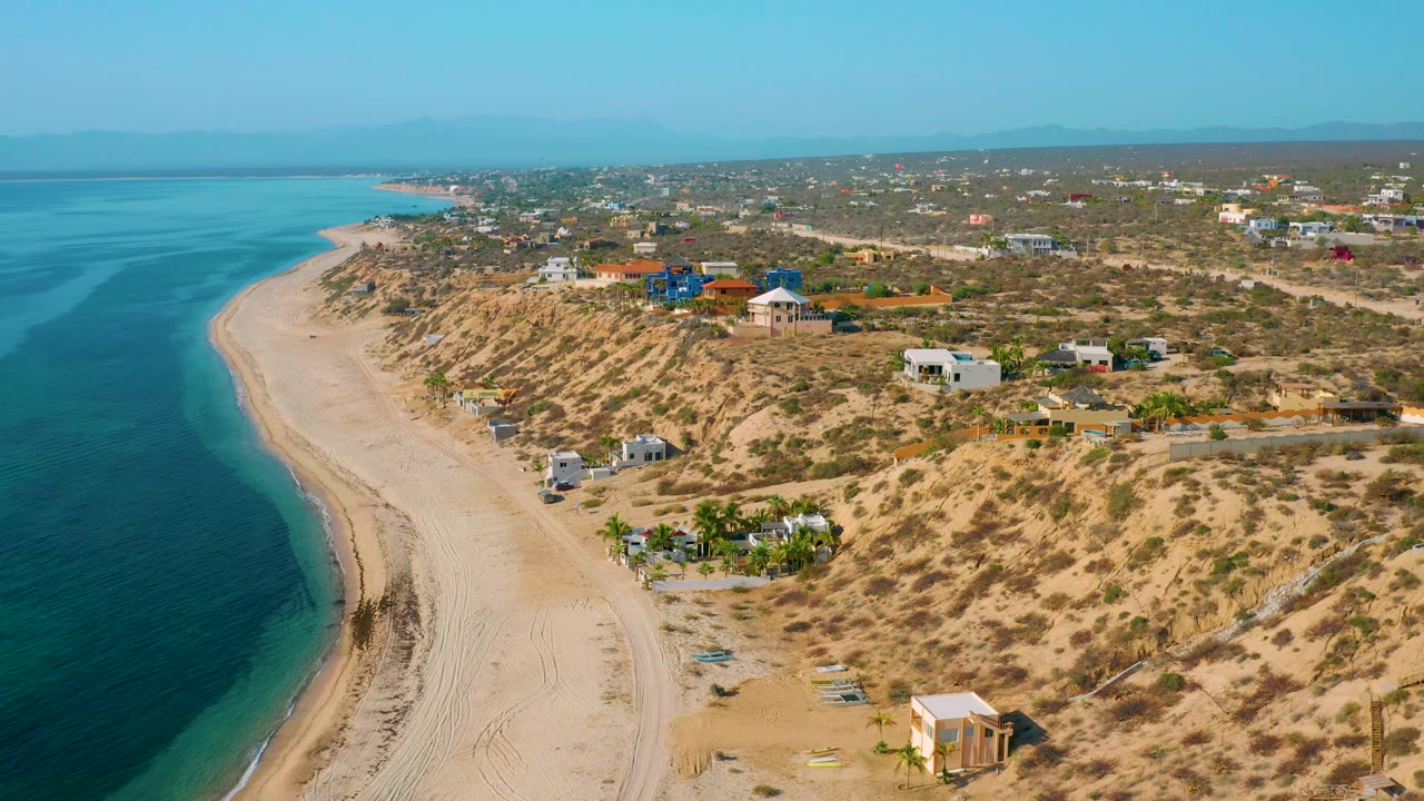 High coastal view with turquoise shallows, sand tracks, dunes, and scattered structures.