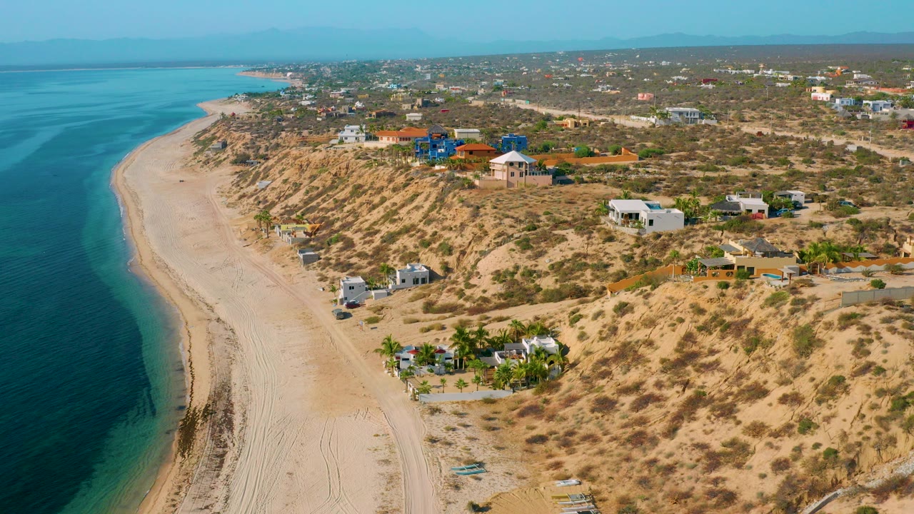 Aerial view over El Sargento La Ventana, Mexico, showing blue water beside a pale curved beach, scrubby dunes, scattered houses, and no visible people.