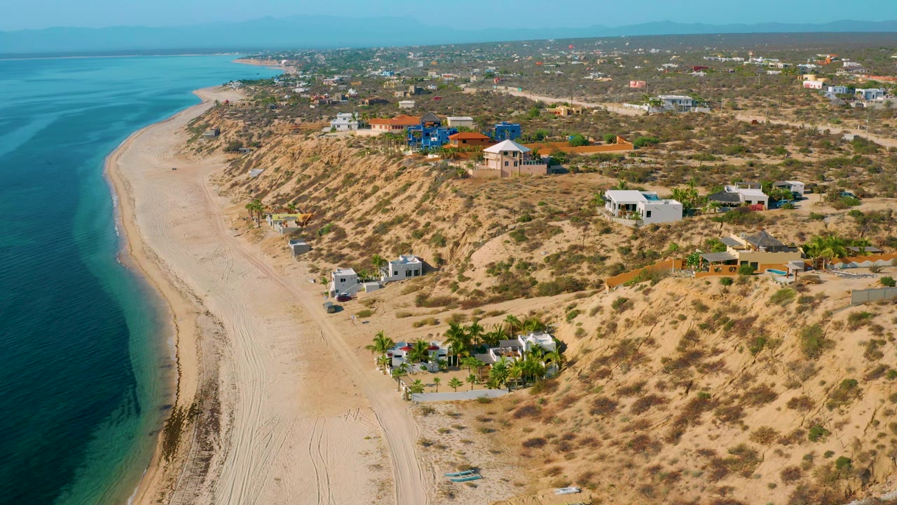 Elevated coastal view over El Sargento La Ventana, Mexico, with the beach running into the distance, homes across dry scrubland, and no visible people.