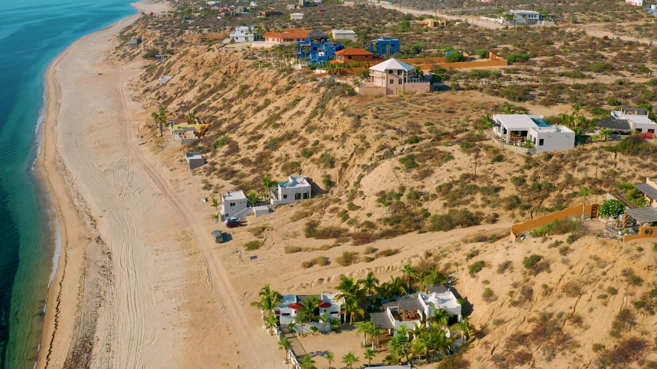 A wide aerial frame shows El Sargento La Ventana's empty beach on the left, with dune vegetation and a cluster of homes on the right, no visible people.