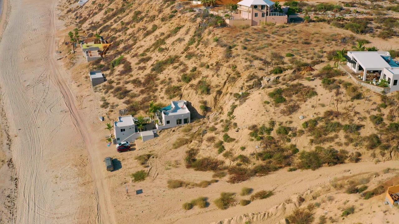 Elevated drone view in El Sargento La Ventana, Mexico, showing a white modern house, parked cars, sandy road, dry hillside scrub, and no visible people.