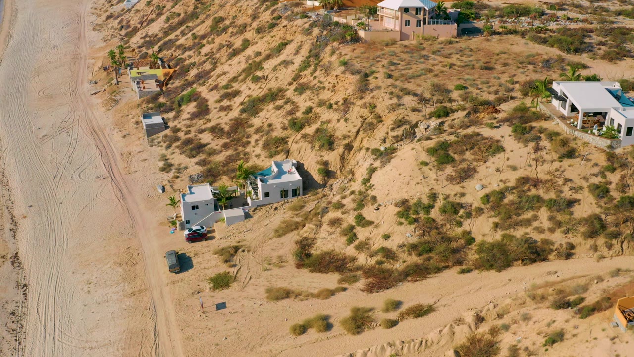 High angled frame over El Sargento La Ventana, Mexico, with the white residence lower left, a sandy road running vertically, hillside brush behind it, and no visible people.