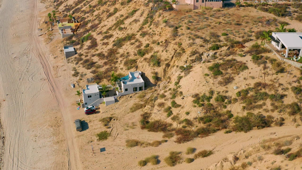 Aerial view in El Sargento La Ventana, Mexico, showing the white house near the bottom, a blue roof or pool area, parked cars, sandy track, scrubland, and no visible people.