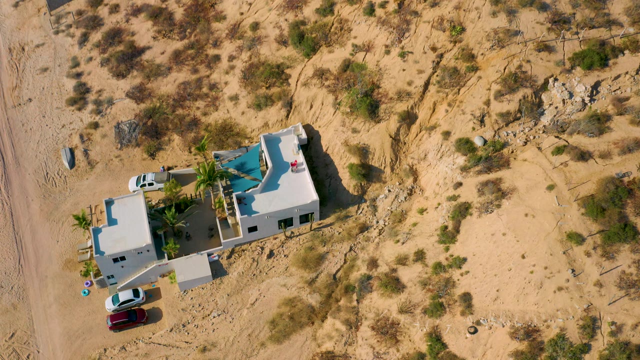 Top-down aerial frame in El Sargento La Ventana, Mexico showing the white villa shifted lower in frame, a white car and red car parked nearby, palms, tire-marked sand, and no clearly identifiable faces.