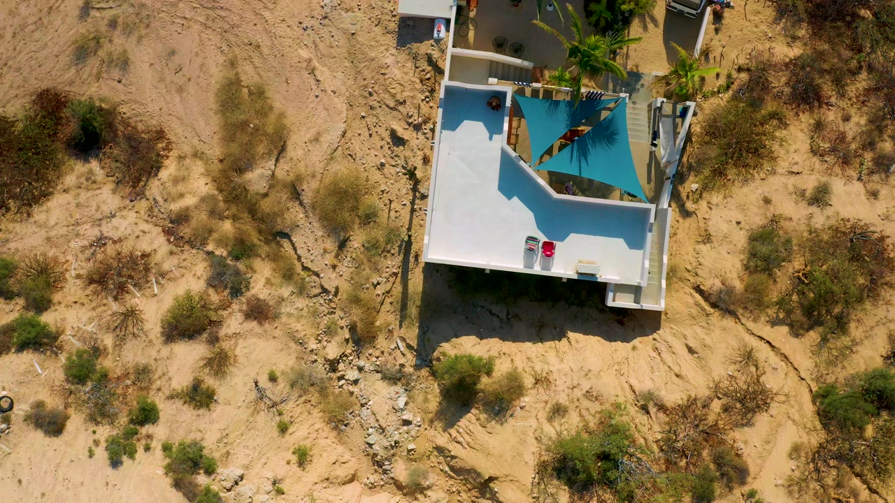 Opening overhead frame in El Sargento La Ventana, Mexico, with a white flat roof, turquoise shade canopy, sandy terrain, scattered bushes, and no visible people.