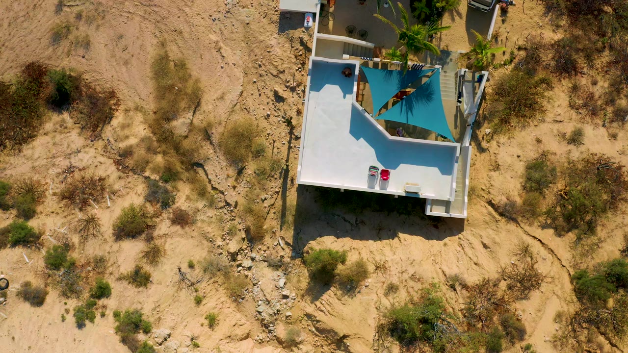 Top-down drone view of a white house roof and surrounding sandy brush.