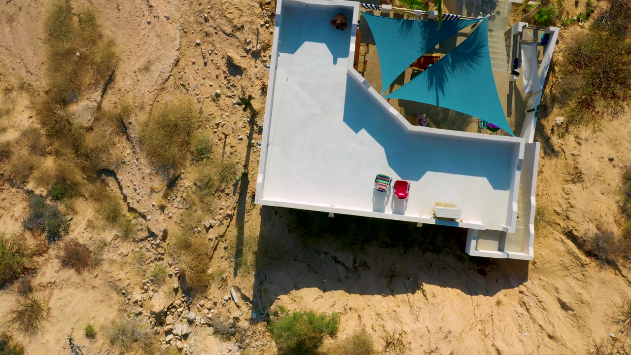 Top-down frame in El Sargento La Ventana, Mexico showing the terrace edge, blue-green shade sail, white building surfaces, dry surrounding ground, and no visible people.