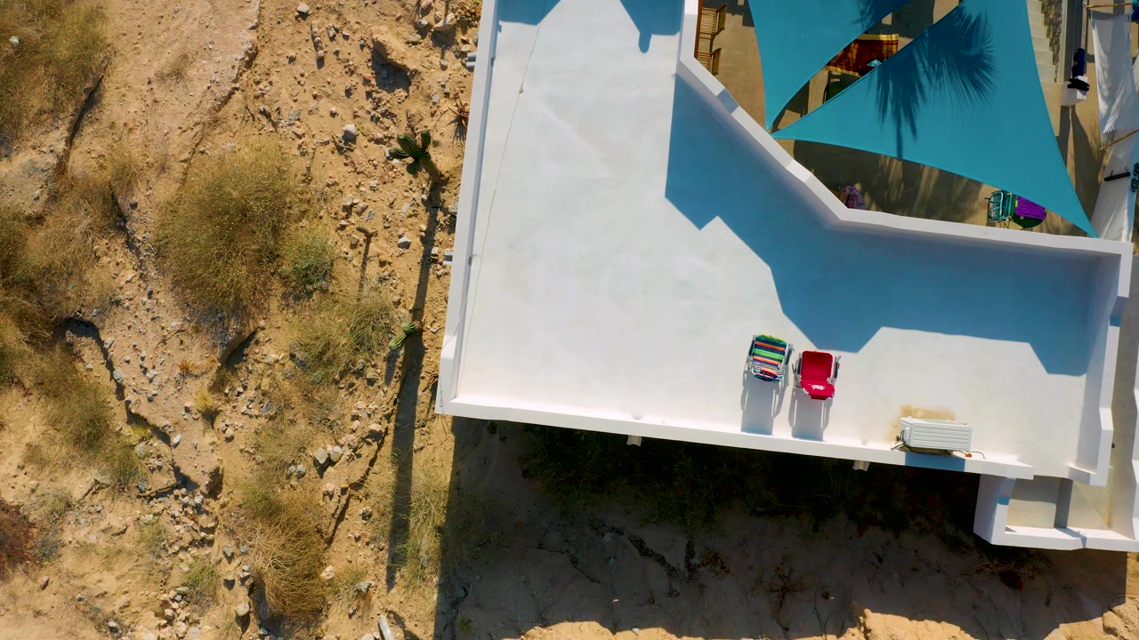Top-down frame in El Sargento La Ventana, Mexico showing a white rooftop terrace with two lounge chairs near the edge, blue shade fabric above, dry rocky ground at left, and no visible people.