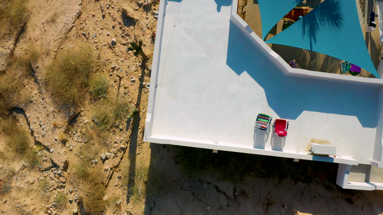 Top-down El Sargento La Ventana, Mexico frame showing two lounge chairs on a bright white terrace, a large angular shadow across the roof, blue shade cloth near the top, and no visible people.