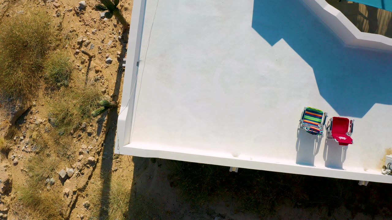 Overhead view in El Sargento La Ventana, Mexico, showing a white flat roof, two outdoor chairs near the lower right edge, sandy rocky ground at left, and no visible people.