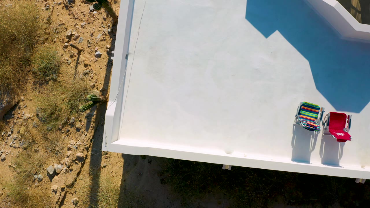 Aerial view in El Sargento La Ventana, Mexico, showing the white rooftop shifted farther across frame, bright chairs near the right edge, tan rocky ground at left, and no visible people.