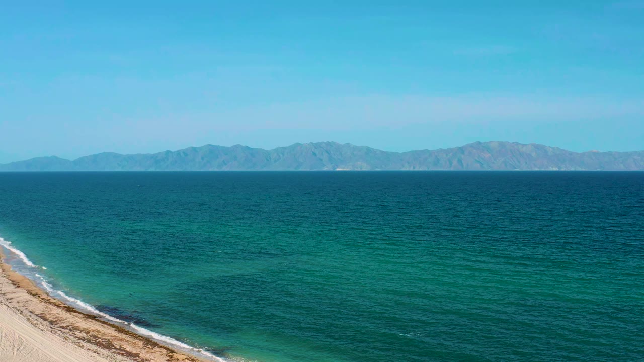 Opening frame over El Sargento La Ventana water with pale sand entering from the lower left, teal shallows, distant mountains on the horizon, and no visible people.