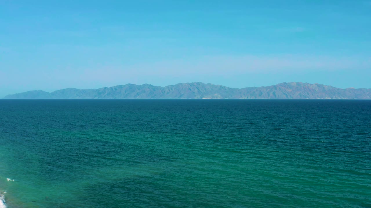 Blue-green sea fills the foreground near El Sargento La Ventana, Mexico, with a long rocky landform across the horizon and no visible people.