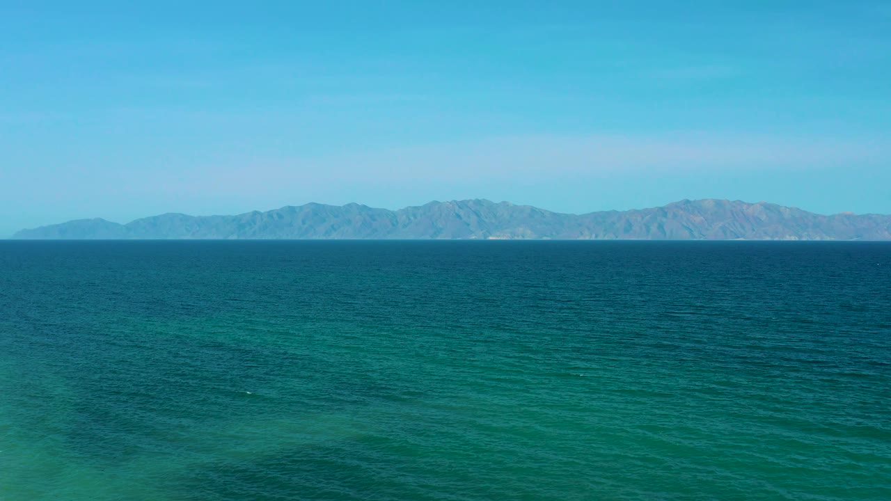 Open ocean sits under a clear sky near El Sargento La Ventana, Mexico, with faint wave marks in the water and mountains spanning the background, no visible people.