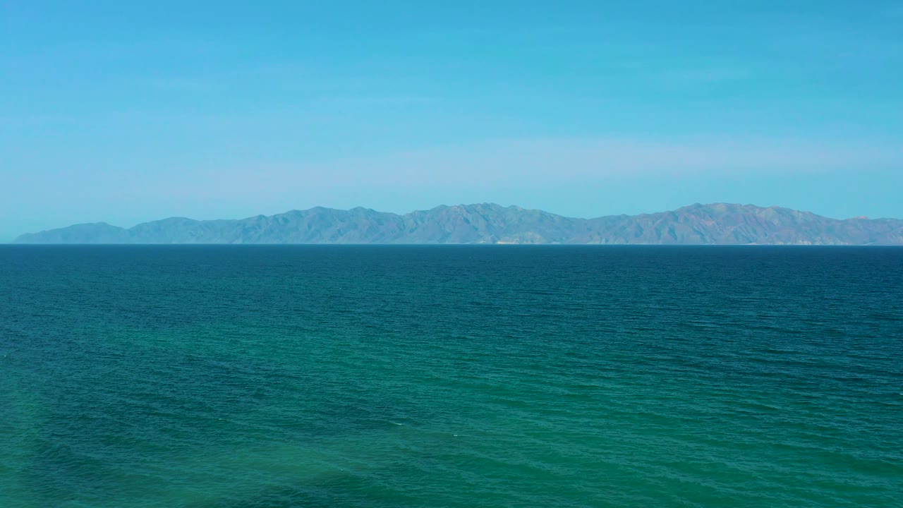 A wide view of green-blue water near El Sargento La Ventana, Mexico, shows darker ocean bands in the midground and low mountains along the horizon, no visible people.