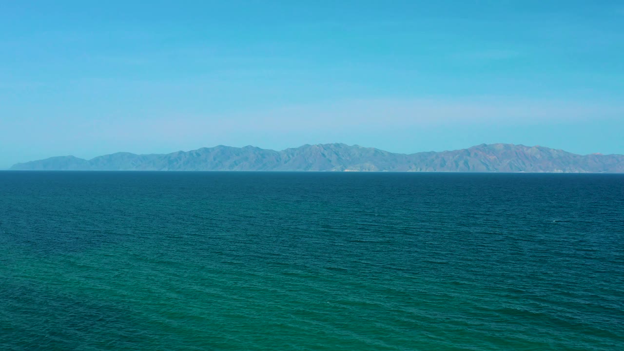 Frame at 0:02 in El Sargento La Ventana, Mexico, showing textured teal water, pale distant mountains, clear sky, and no visible people.