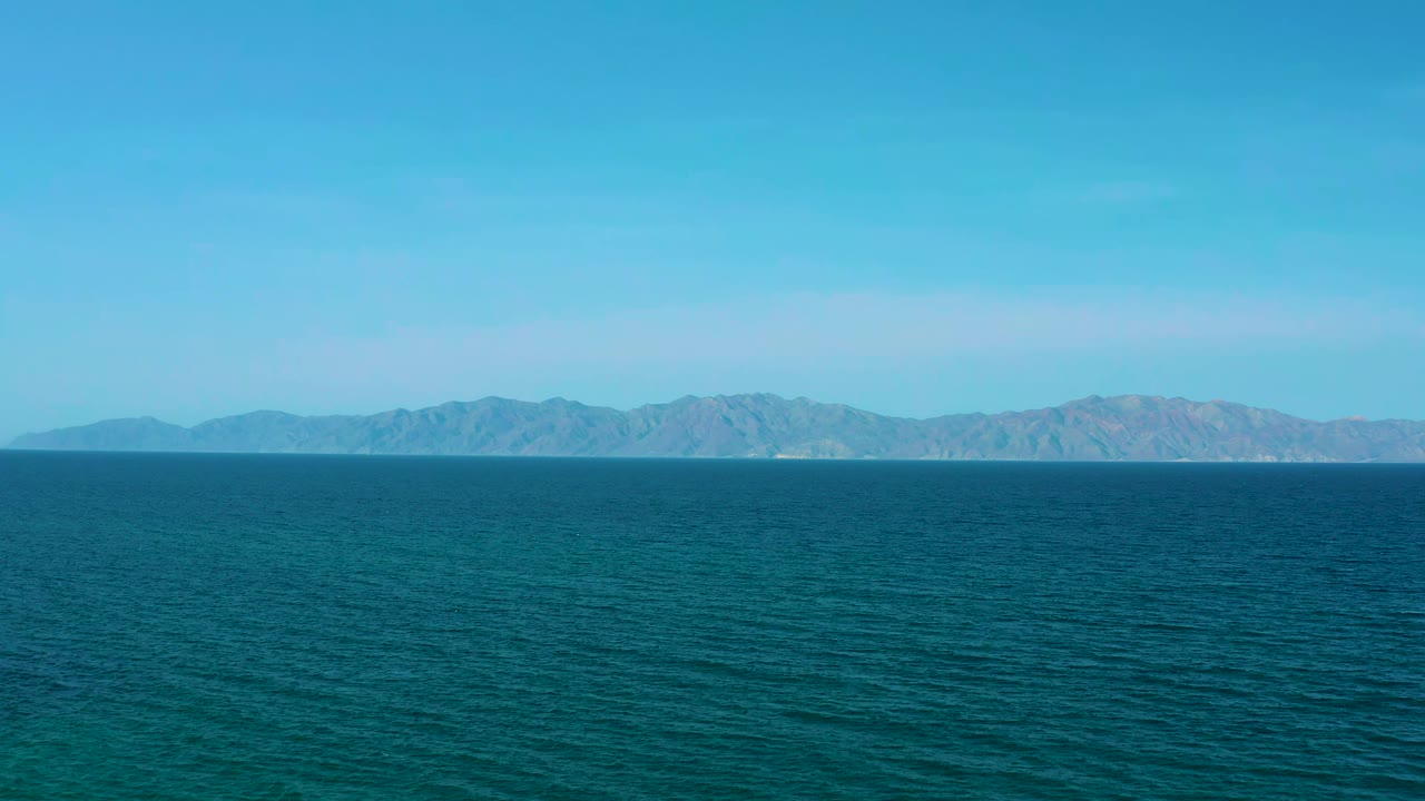 El Sargento La Ventana coastal frame with rippled blue water in front, a long distant landform across the center, pale sky above, and no visible people.