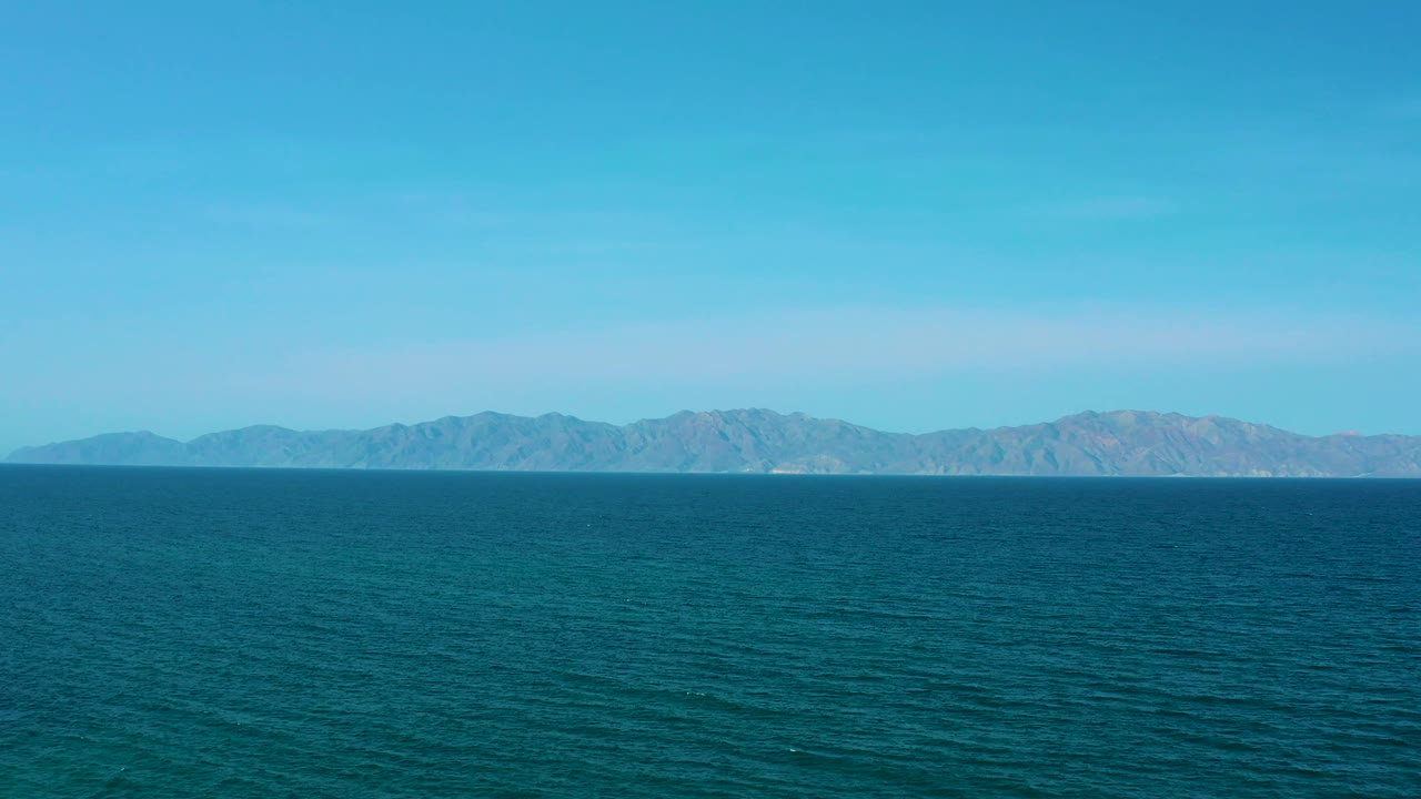 Wide 4K frame from El Sargento La Ventana showing darker teal water below, a hazy island ridge across the horizon, clean daylight sky, and no visible people.