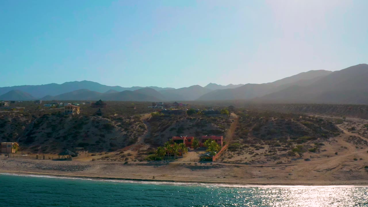 A wide aerial view in El Sargento La Ventana, Mexico shows sun glitter on the water, a thin beach strip, low structures, dry coastal land, distant mountains, and no visible people.