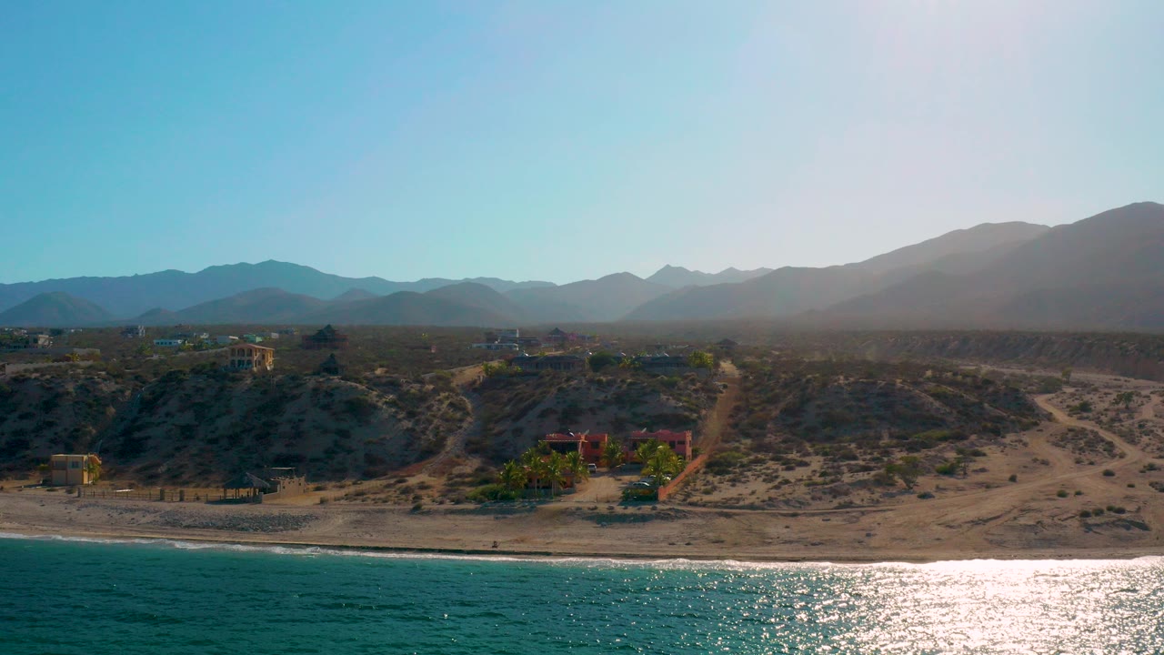 Wide aerial frame over El Sargento La Ventana, Mexico, with turquoise sea in the foreground, a bright water reflection, sandy shoreline, scattered buildings, hazy mountains, and no visible people.