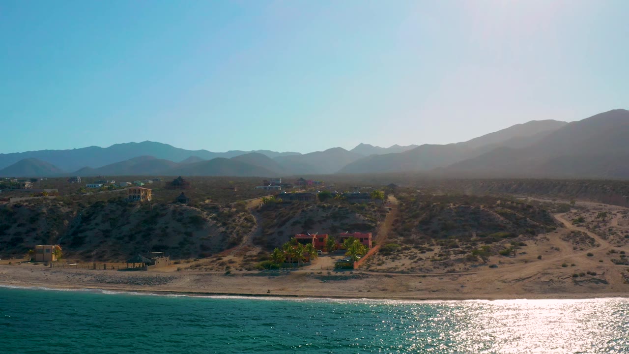 El Sargento La Ventana coastal frame with turquoise water at the bottom, a pale sand strip, sparse buildings across dry terrain, layered mountains in haze, and no visible people.
