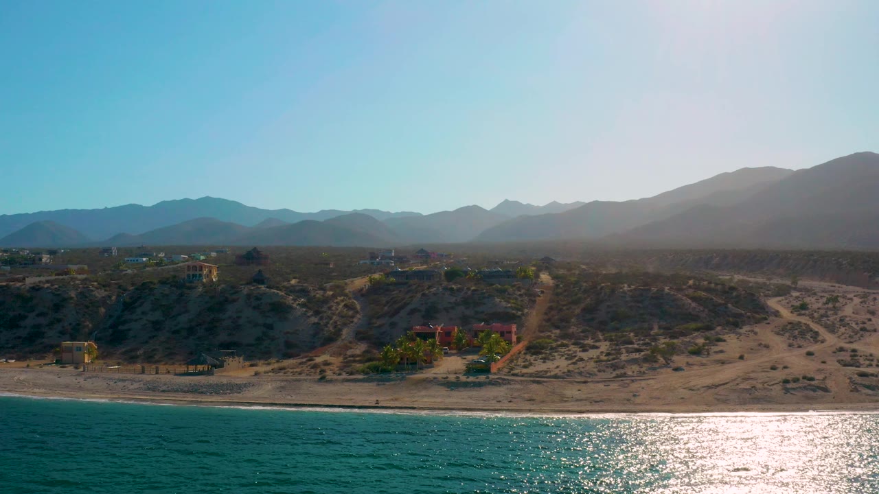 Sunlit shoreline with sea in front, desert lots, houses, and mountains behind