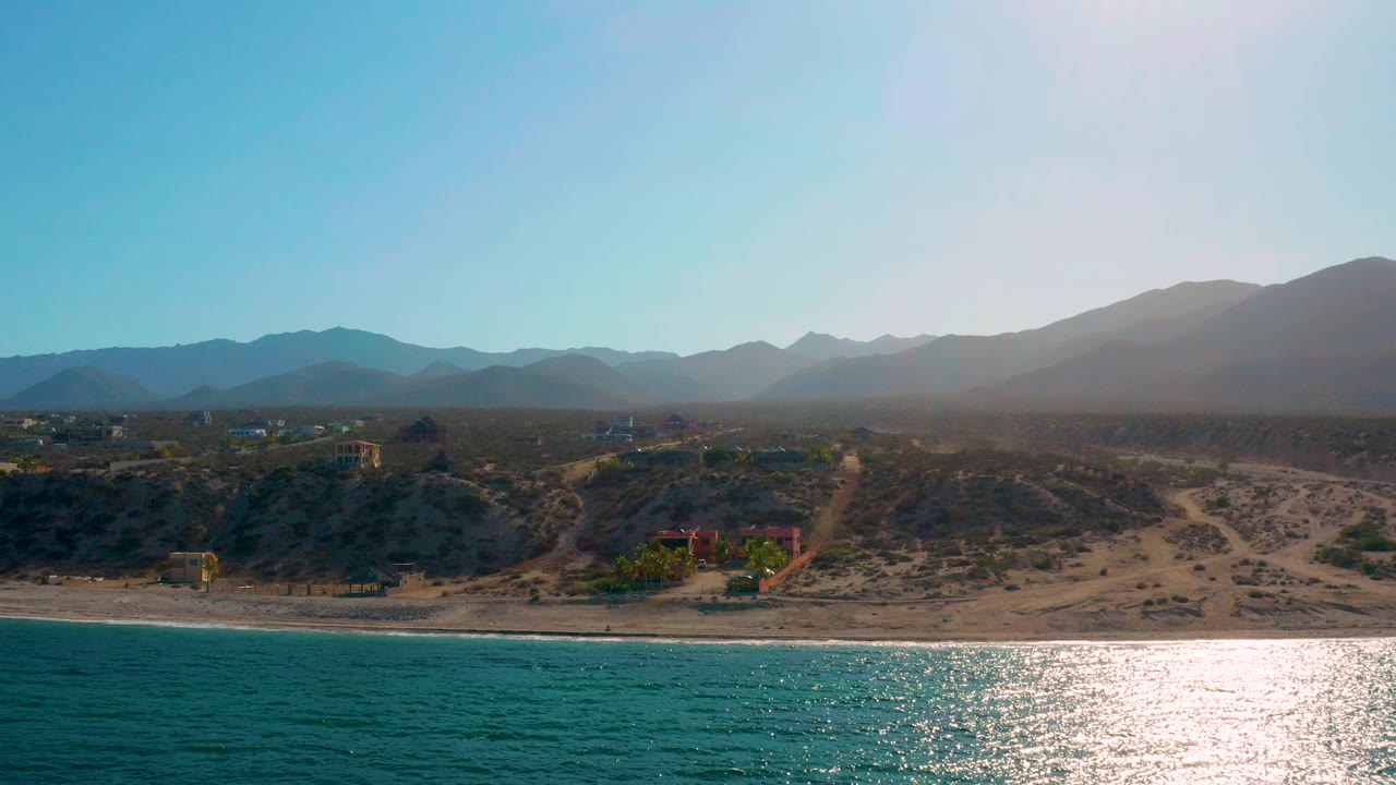 Opening aerial frame over El Sargento La Ventana, Mexico, with turquoise water at the bottom, sun glare near shore, sandy dunes, scattered inland buildings, hazy mountains, and no visible people.