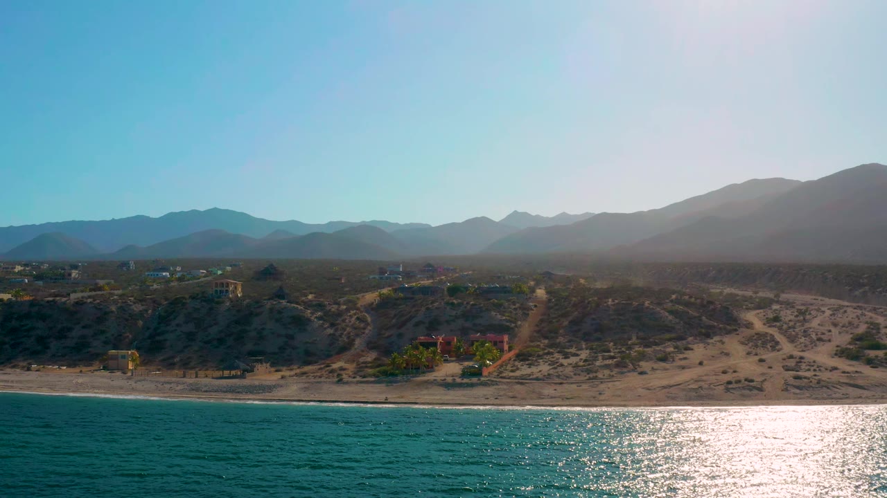 Coastal aerial with bright water reflection, dunes, scrub, and mountain horizon