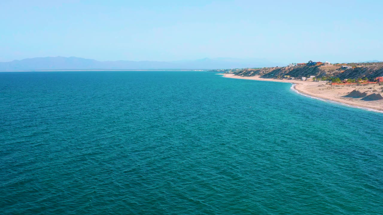 Opening aerial frame over El Sargento La Ventana, Mexico, with turquoise water in the foreground, a sandy shore at right, hazy mountains on the horizon, and no visible people.