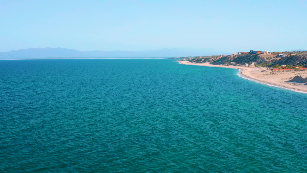 Wide aerial view over blue-green ocean at El Sargento La Ventana, Mexico, with sandy shoreline and low hills on the right, distant mountains on the horizon, and no visible people.