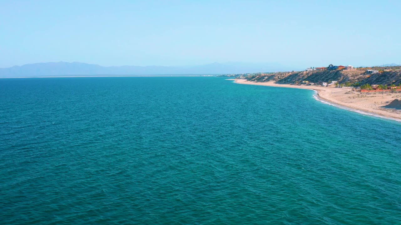 Blue ocean fills the foreground and midground at El Sargento La Ventana, Mexico, with a pale beach and low developed hillside to the right, distant mountains under clear sky, and no visible people.