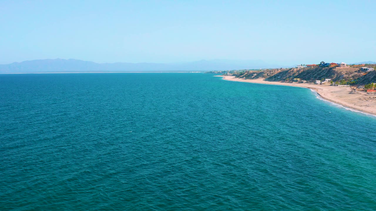 Aerial view over blue green water toward the tan shoreline and bluff at El Sargento La Ventana, Mexico, with hazy land on the horizon and no visible people.
