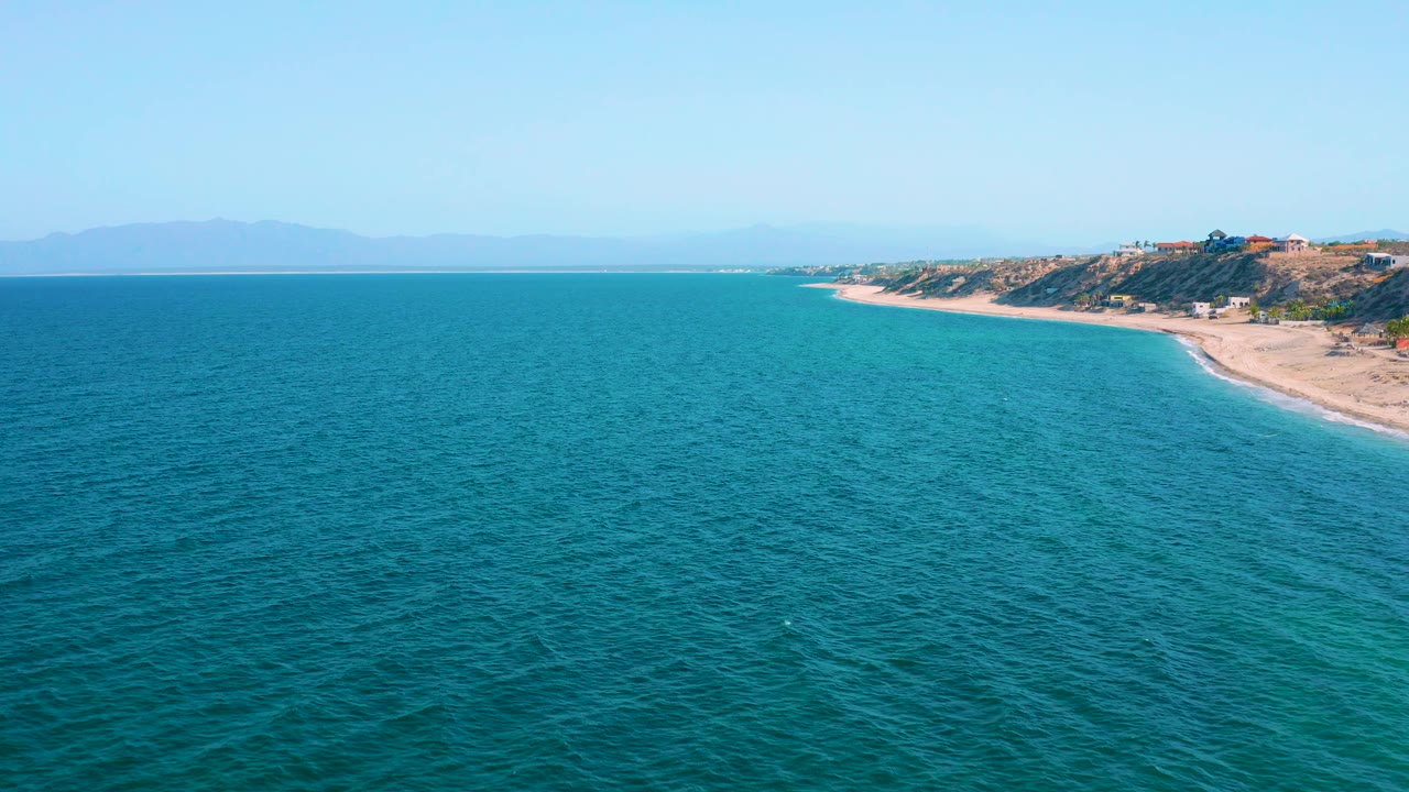 Aerial coastal view at El Sargento La Ventana, Mexico, with rippled blue water across the foreground, pale tan beach at right, bluff-top buildings, and no visible people.