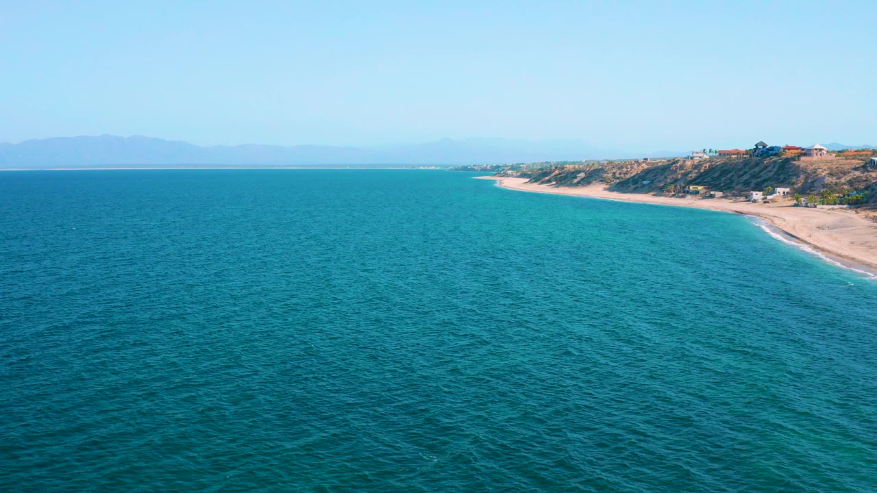 High aerial view in El Sargento La Ventana, Mexico, with blue ocean filling the foreground, a curving empty beach on the right, hazy land on the horizon, and no visible people.