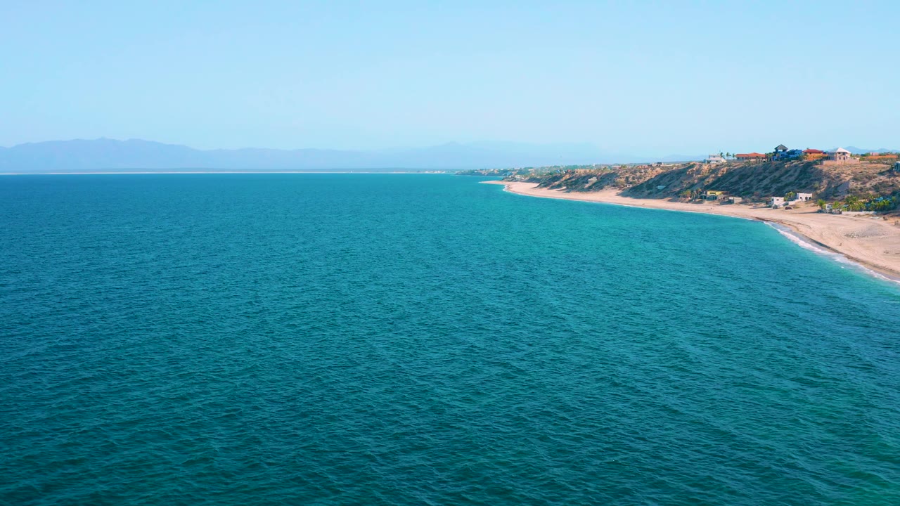 Wide high view in El Sargento La Ventana, Mexico, with open blue water across most of the frame, a narrow empty beach and coastal bluffs near the upper right, and no visible people.