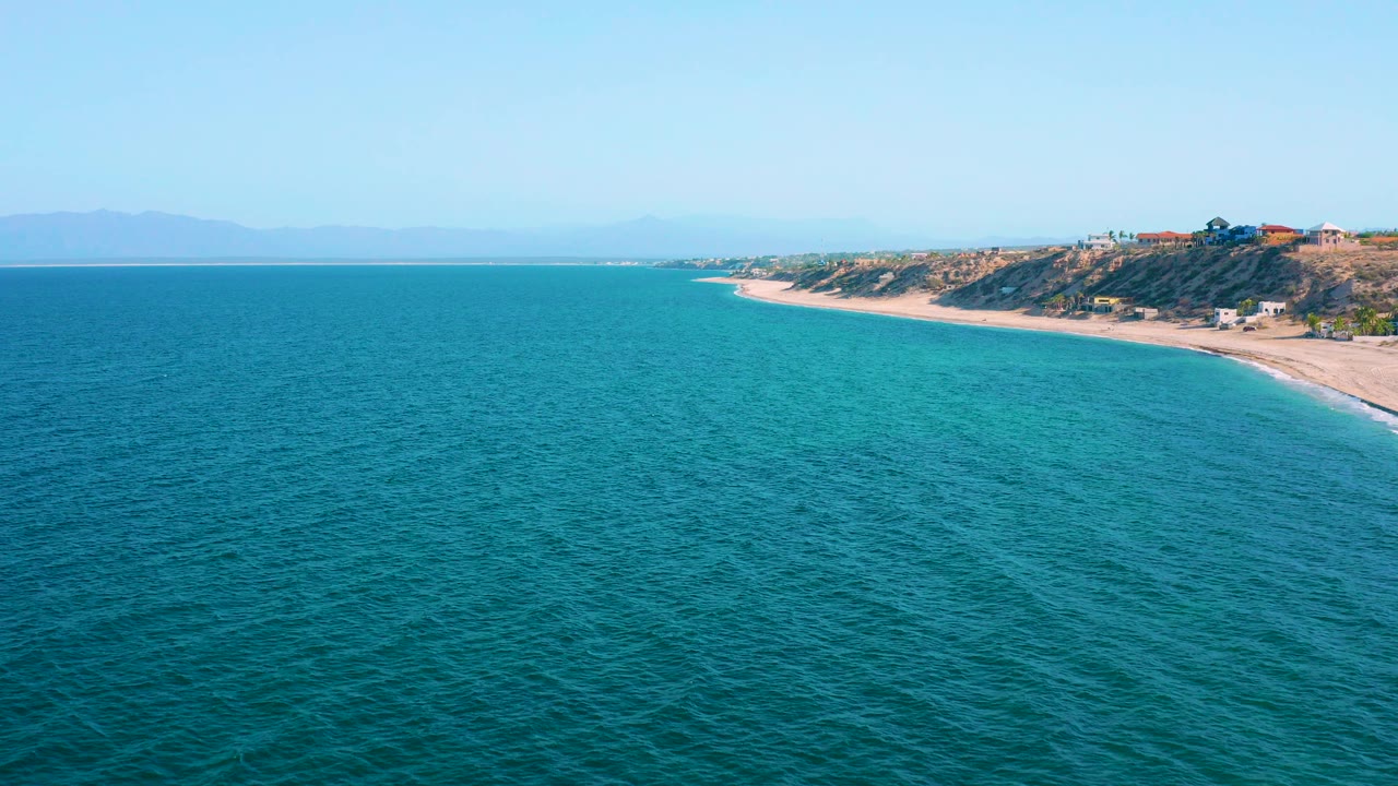 Aerial view in El Sargento La Ventana, Mexico, with deep blue ocean in the foreground, pale beach and low bluffs on the right, hazy mountains on the horizon, and no visible people.