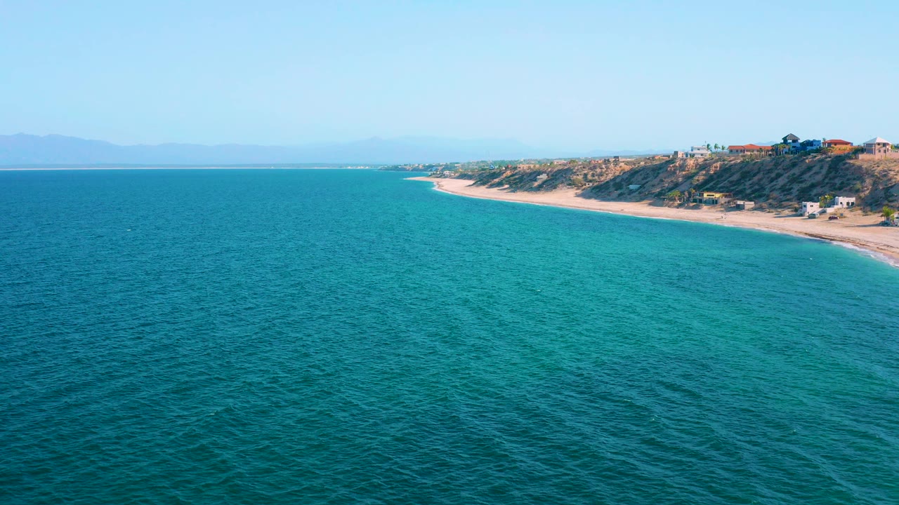 Aerial view over blue ocean toward the sandy El Sargento La Ventana shoreline, with dunes, shrubs, scattered houses, distant hills, and no visible people.