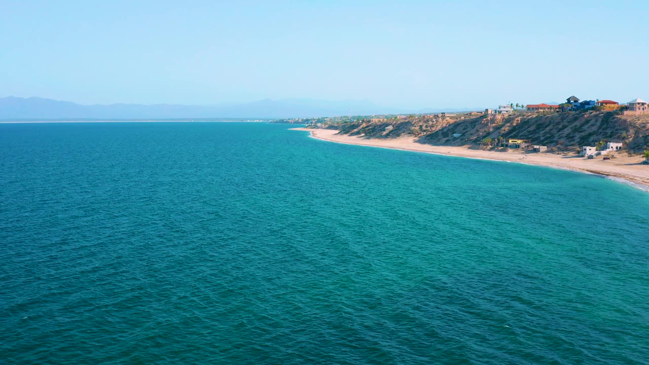 Wide aerial frame of the El Sargento La Ventana coastline, showing turquoise water shifting to deeper blue, a narrow sand beach, scrub-covered dunes, hillside houses, and no visible people.