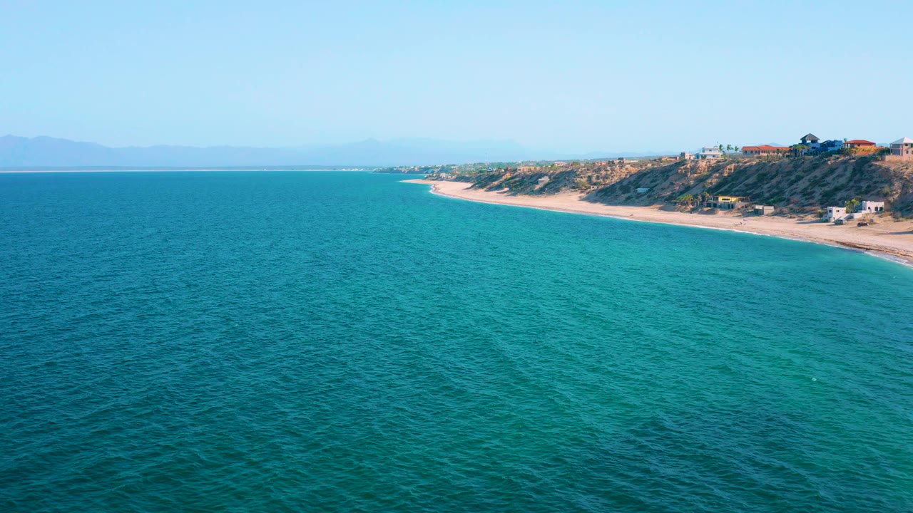 Aerial ocean view with a curved sandy beach, dune ridge, shrubs, and shoreline houses.
