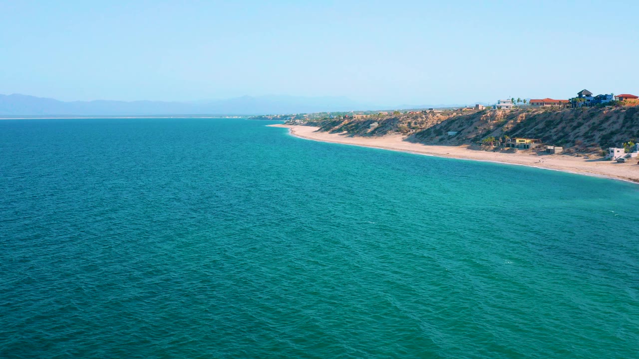 Opening aerial view in El Sargento La Ventana, Mexico, with turquoise water in the foreground, a pale beach curving along a scrubby bluff, distant haze, and no visible people.