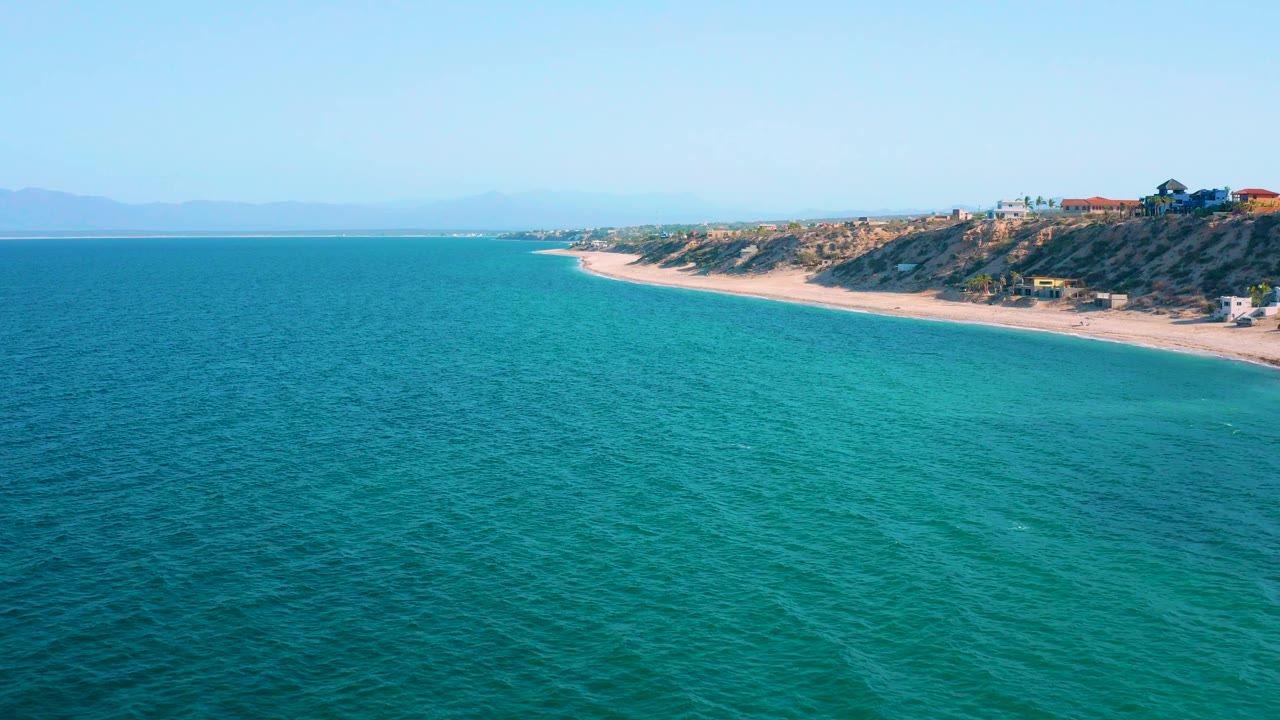 Aerial coastal frame from El Sargento La Ventana, Mexico, showing rippled blue-green water, a light sand shoreline, sparse hillside buildings, hazy mountains, and no visible people.