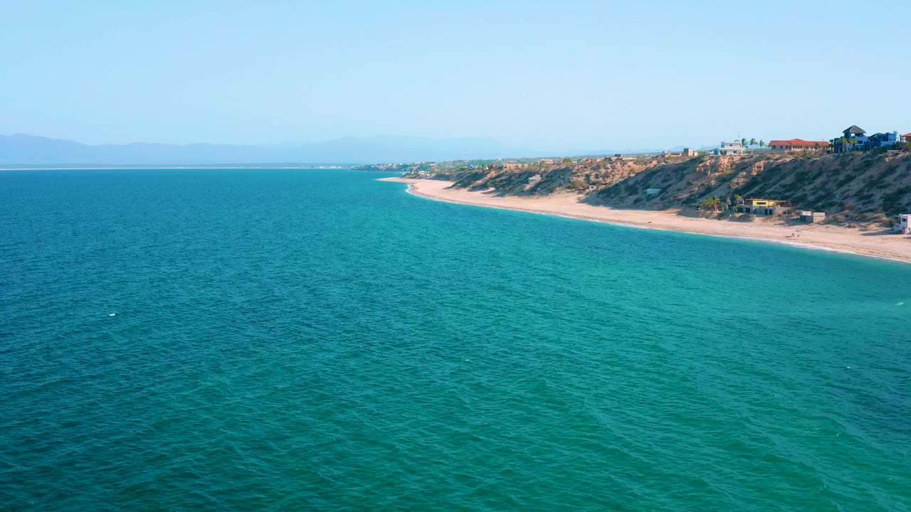 Coastal aerial view with turquoise sea, sandy beach, and scrub-covered bluff.