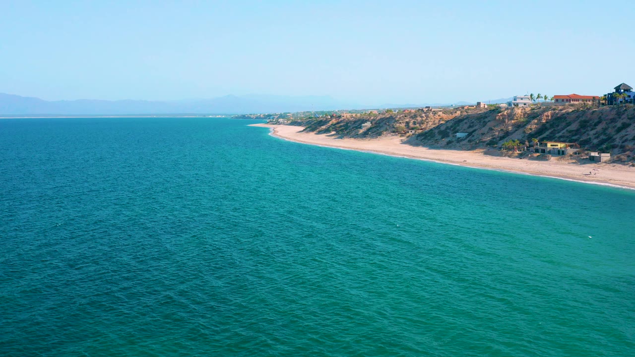 Opening frame of El Sargento La Ventana with turquoise water in the foreground, a pale beach curving along low bluffs, distant mountains on the horizon, and no visible people.