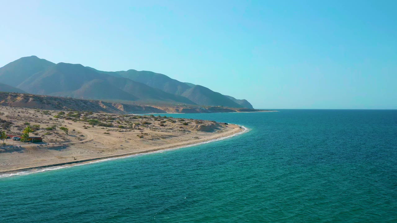 Wide aerial frame of El Sargento La Ventana, Mexico, showing blue-green water meeting a narrow pale beach, dry coastal land, layered mountains, and no visible people.