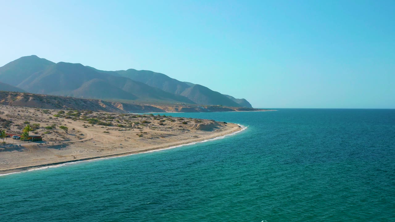 Aerial shoreline with turquoise water, narrow surf line, dry beach, and mountains.